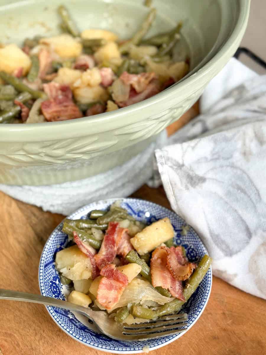 A small blue and white bowl contains green beans, potatoes, onions, and slices of bacon. The same dish is in a larger green serving bowl in the background, with a fork and white patterned napkin nearby.