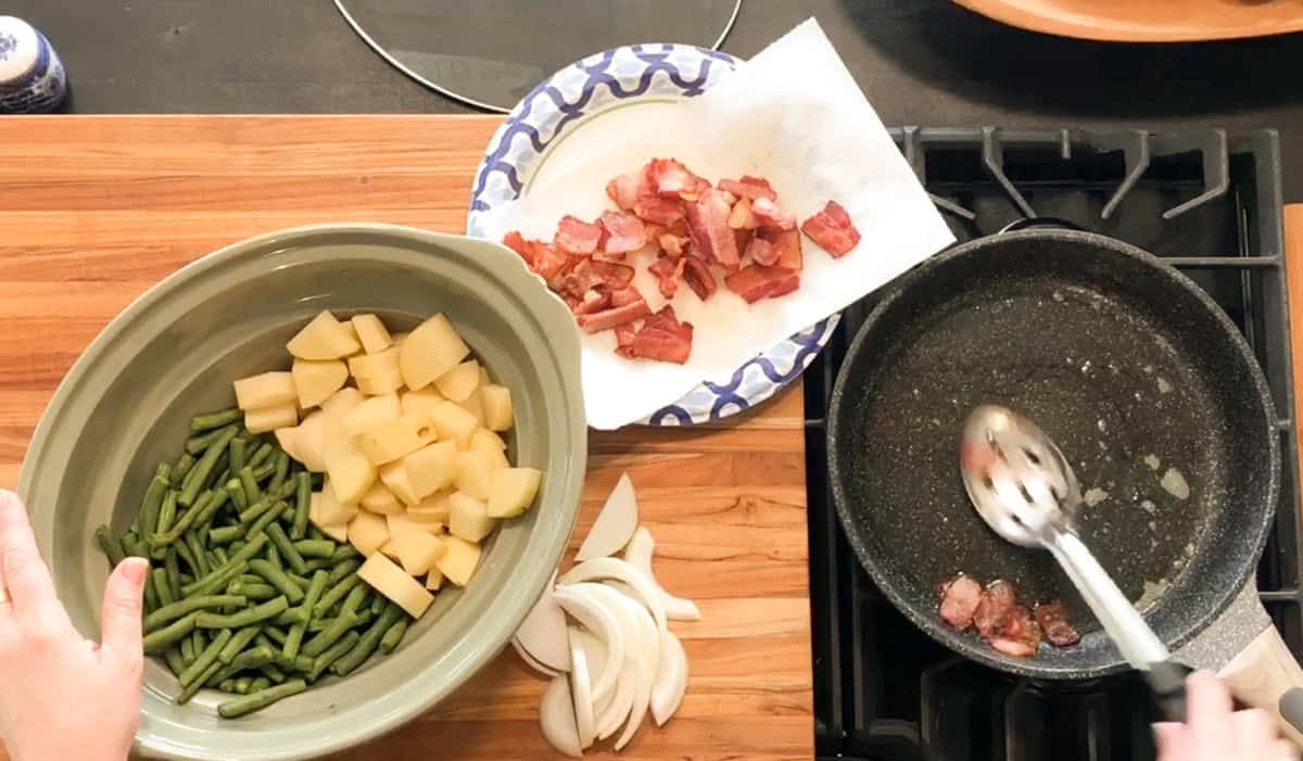 A person uses a slotted spoon to remove cooked bacon from a frying pan; nearby are a bowl with green beans and diced potatoes, sliced onions, and a plate with bacon pieces on a paper towel.