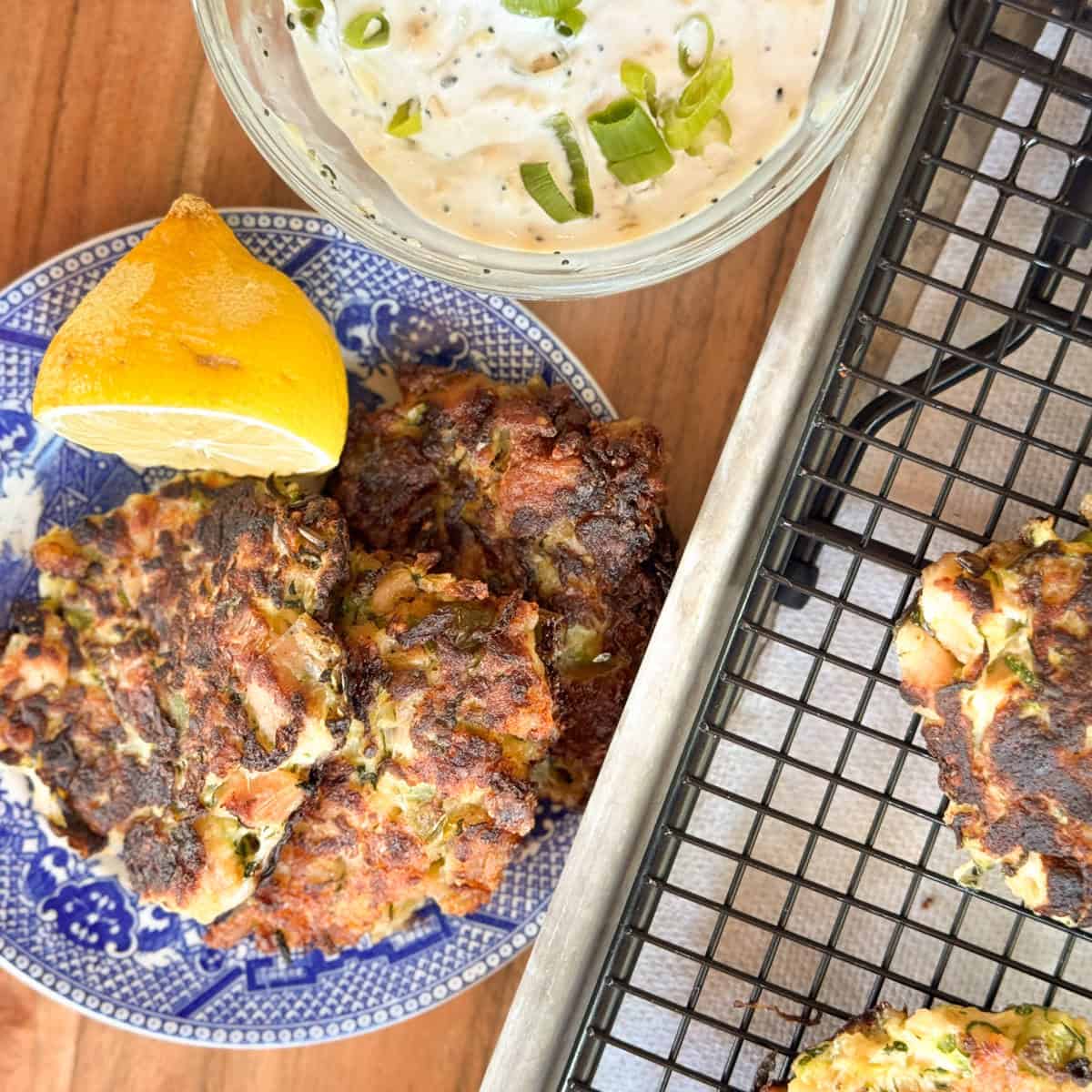 Two vegetable fritters on a blue plate with a lemon wedge, a bowl of creamy dipping sauce with green onions, and additional fritters on a cooling rack on a wooden surface.
