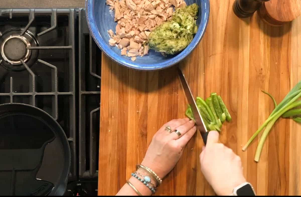 A person slices green onions on a wooden cutting board next to a blue bowl containing chopped tuna and mashed avocado, with a stove and skillet visible to the left.