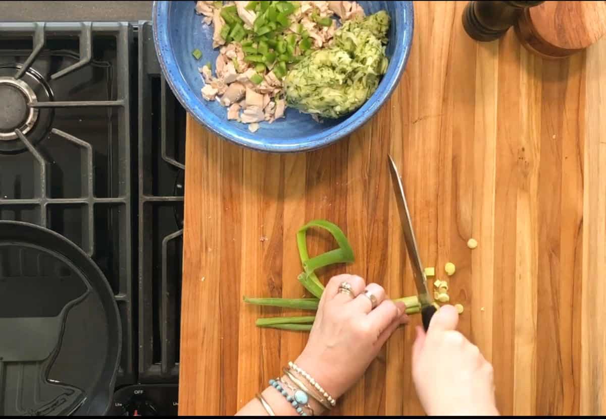 A person’s hands chop green onions on a wooden cutting board next to a stove. A blue bowl with chopped vegetables and possibly chicken is nearby, along with a knife and a pepper grinder.