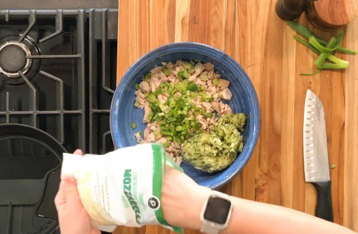 A person holds a bag of shredded mozzarella cheese above a blue bowl containing chopped chicken, green onions, and shredded zucchini on a wooden countertop beside a knife and green onions.