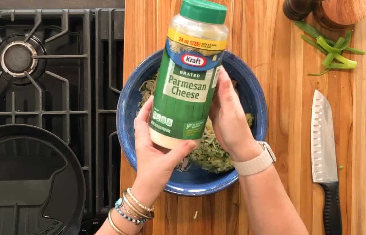 A person holds a container of Kraft grated Parmesan cheese over a blue bowl on a wooden countertop with a knife, sliced green pepper, and a black stove visible nearby.