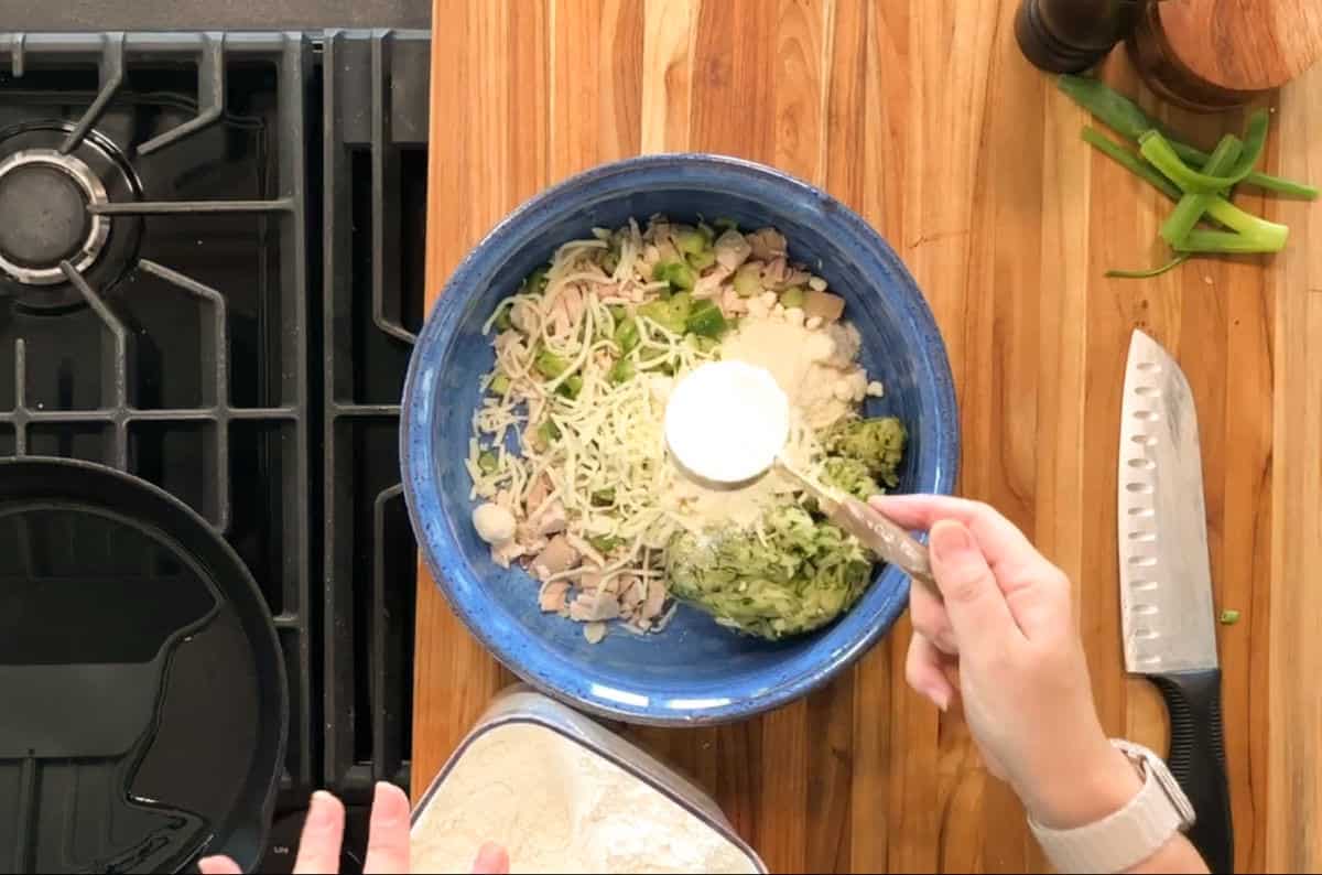A person holds a measuring cup of flour over a blue mixing bowl containing shredded cheese, chopped chicken, green onions, and other ingredients on a wooden counter beside a stove. A knife and sliced green onions are nearby.
