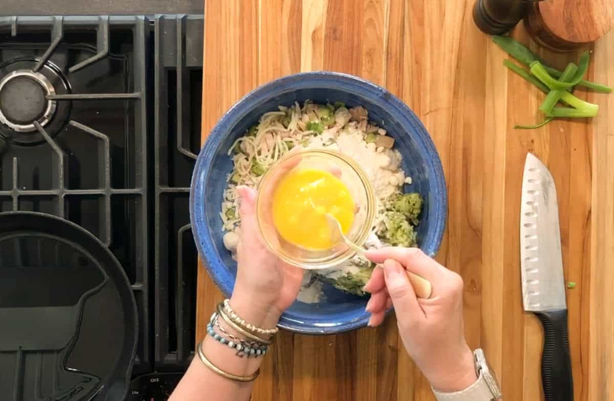 A person holds a small bowl of beaten eggs above a blue bowl containing chopped vegetables and other ingredients on a wooden countertop next to a stove, knife, pepper mill, and sliced green onions.