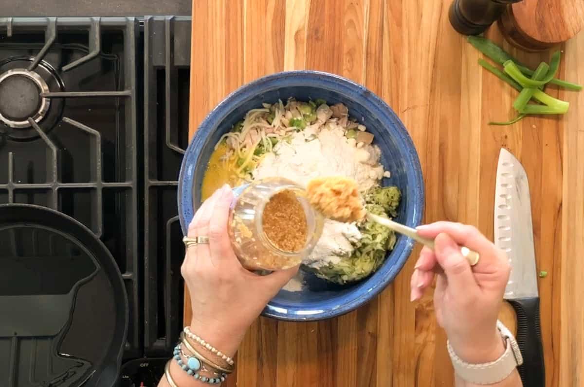 A person adds a spoonful of a brown granular ingredient from a small jar to a blue bowl filled with flour, eggs, and vegetables on a wooden countertop beside a stove and a chopping knife.
