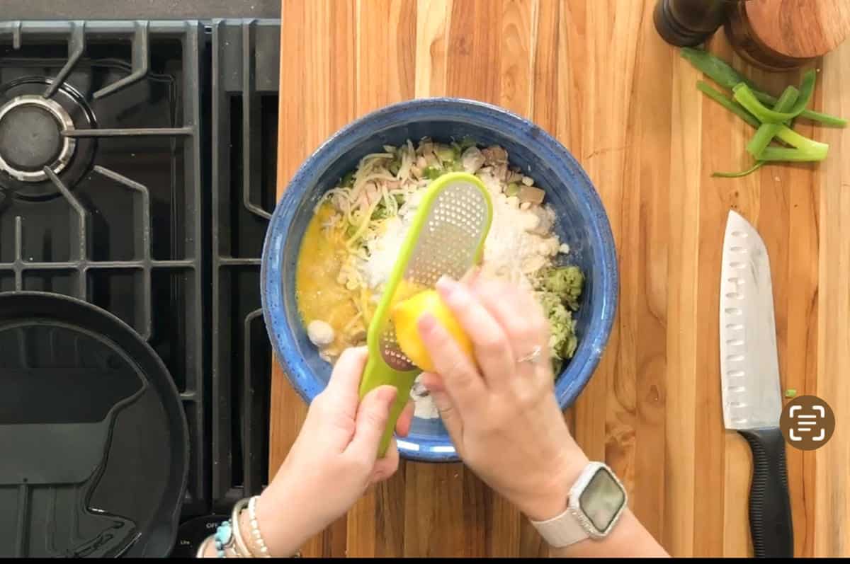 A person grates cheese into a blue bowl containing eggs, vegetables, and shredded meat on a wooden countertop. A knife, green pepper slices, and a pepper grinder are nearby. A stove and skillet are visible on the left.