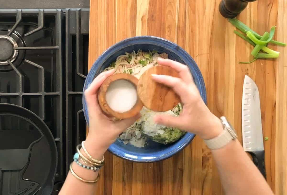 A person sprinkles salt from a wooden container into a blue mixing bowl filled with chopped vegetables on a wooden countertop next to a knife, green onions, a pepper grinder, and a stovetop.