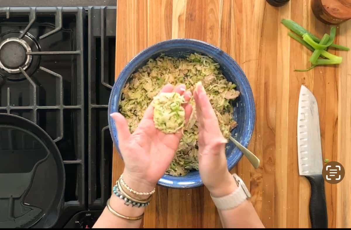 A person shapes a mixture in their hands over a wooden counter with a blue bowl, chef’s knife, green onions, and a stove with a skillet nearby. The mixture appears to contain shredded vegetables.