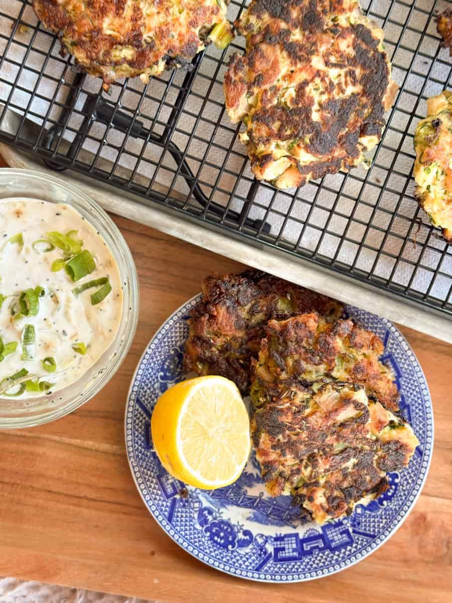 Two cooked vegetable fritters are on a blue patterned plate with a halved lemon. More fritters are on a cooling rack above. A small bowl of creamy dipping sauce topped with chopped green onions is on the left.