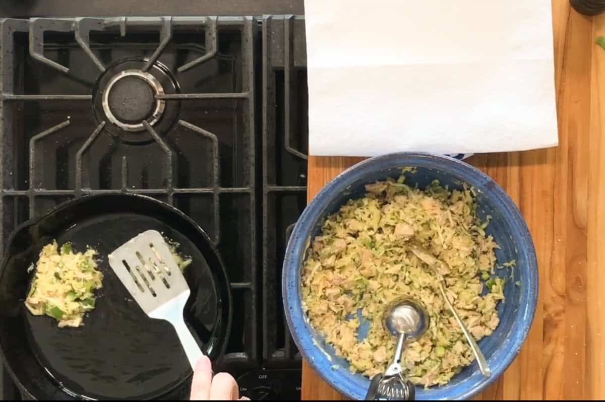 A hand uses a spatula to cook a patty in a skillet on a gas stove, while a blue bowl filled with a chunky mixture and a metal scoop sits nearby on a wooden countertop with paper towels.