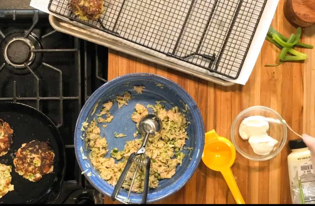 Overhead view of a kitchen counter with a blue bowl containing a mixture and a metal scoop, a cooling rack on a baking sheet, a measuring cup, a small bowl of white sauce, green chili peppers, and a stove with food frying.