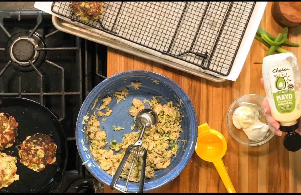A bowl of mixed ingredients and an ice cream scoop sit on a wooden counter next to a yellow measuring cup, a wire rack with food, a stove with patties cooking, and a hand holding a bottle of avocado oil mayo over a small bowl.