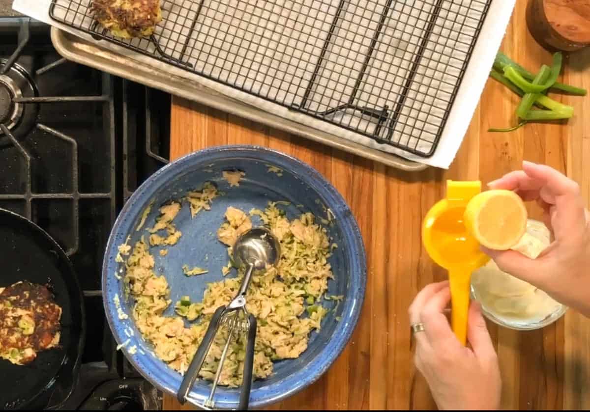 A person squeezes a lemon into a clear glass with a yellow juicer next to a blue bowl of tuna mixture, a metal scoop, a cooling rack, and a pan with a patty on a stovetop.