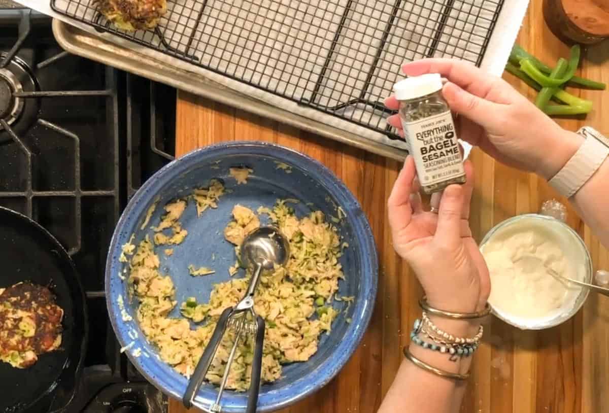 A person holds a jar of everything bagel sesame seasoning above a wooden counter with a mixing bowl, metal scoop, wire rack, and various food ingredients visible.
