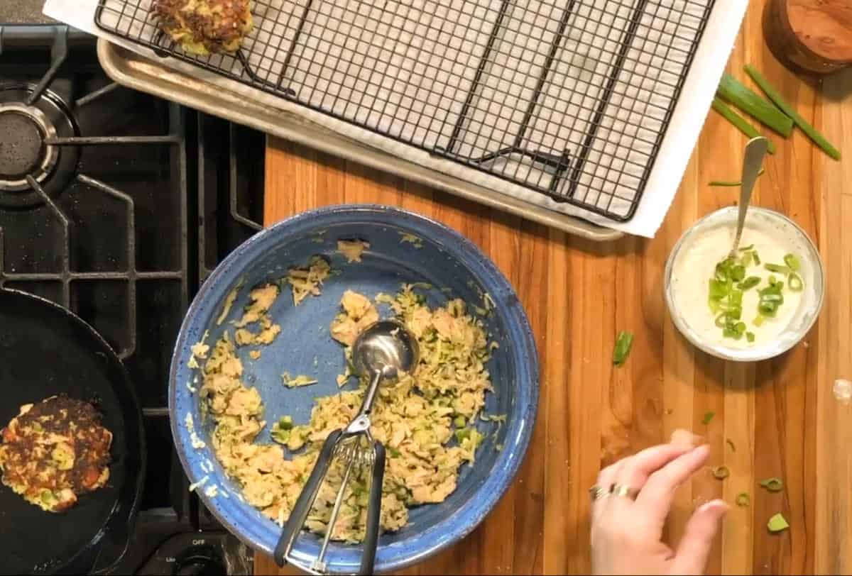 Overhead view of a kitchen scene with a blue bowl of vegetable mixture, a metal scoop, a cooling rack on a tray, a skillet with a patty cooking, a bowl of sauce with chopped green onions, and a hand reaching to grab ingredients.