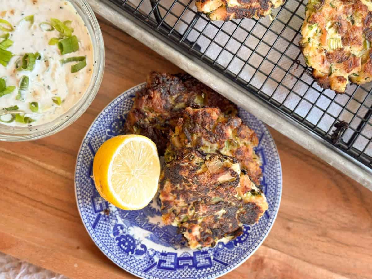 Three browned vegetable fritters are on a blue and white plate with a lemon half. A cooling rack with more fritters and a bowl of creamy dip topped with chopped green onions are nearby on a wooden surface.