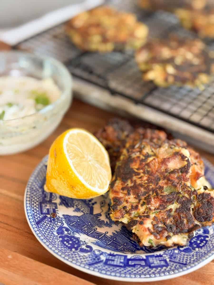 A blue and white plate holds two browned fritters and a lemon half. In the background, more fritters cool on a wire rack, and a glass bowl contains a creamy dip with green herbs.