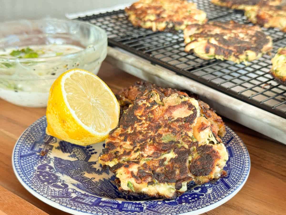 Two vegetable fritters on a blue patterned plate with a halved lemon beside them. A wire rack with more fritters and a bowl of dipping sauce are in the background on a wooden surface.