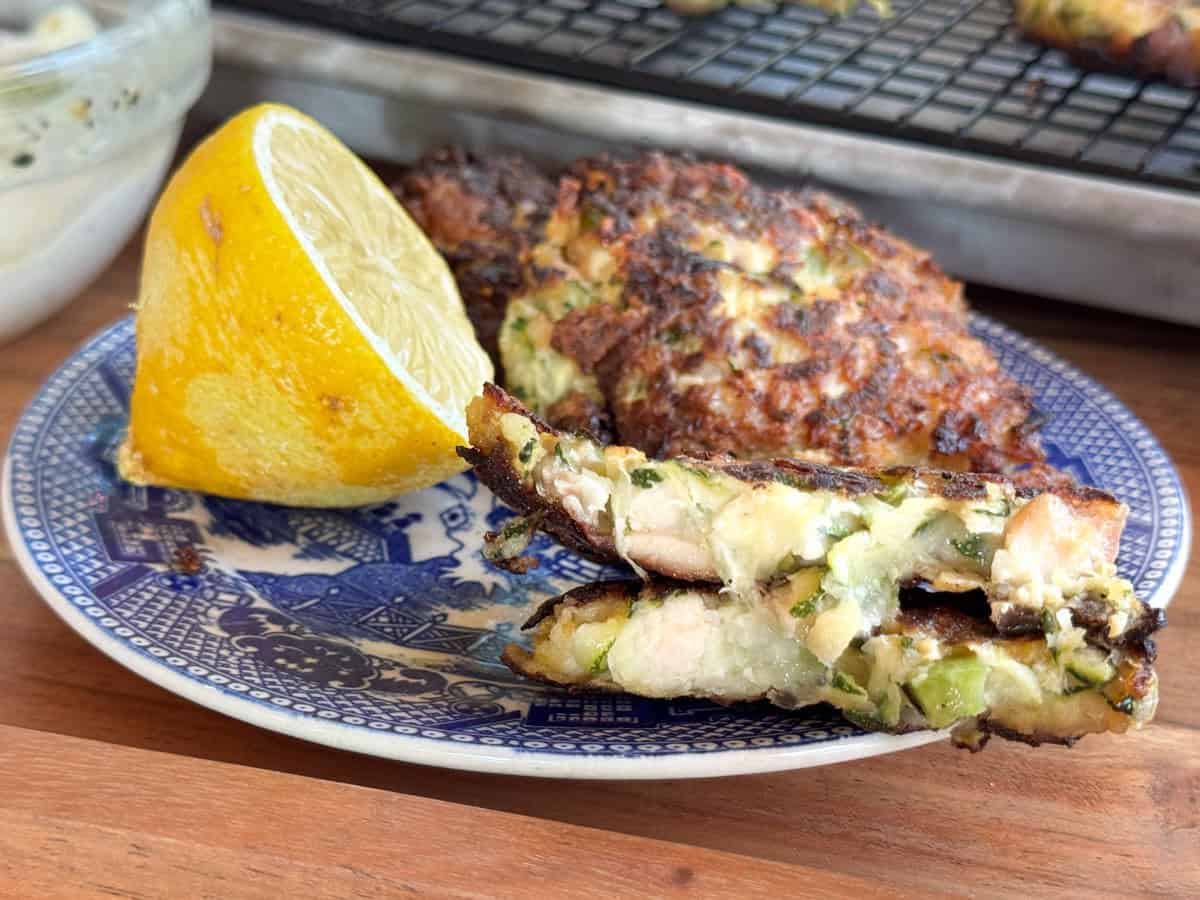 A blue and white plate holds a halved lemon, two zucchini fritters with visible chunks of chicken and herbs, and a whole fritter in the background with a baking tray and cooling rack behind.