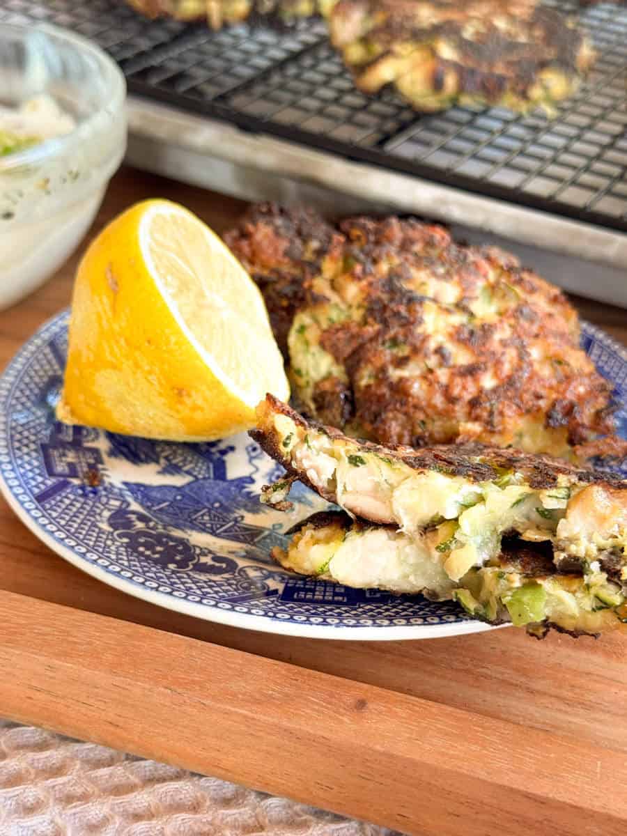 A close-up of a blue patterned plate with vegetable fritters, a lemon wedge, and a small bowl of dip. More fritters are visible on a cooling rack in the background. The fritters appear crispy and browned.
