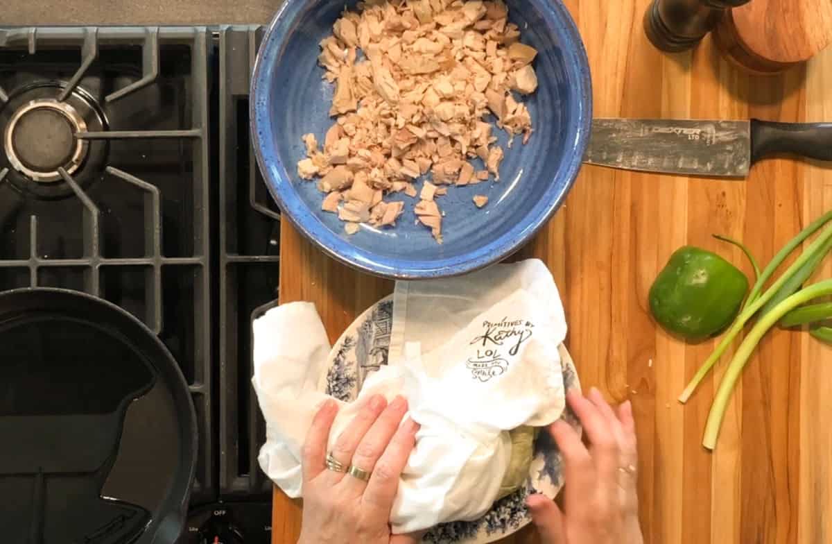 A person presses a paper towel over a plate on a wooden counter beside a large bowl of chopped chicken, a knife, a green bell pepper, and two green onions. Part of a stovetop with a skillet is visible on the left.