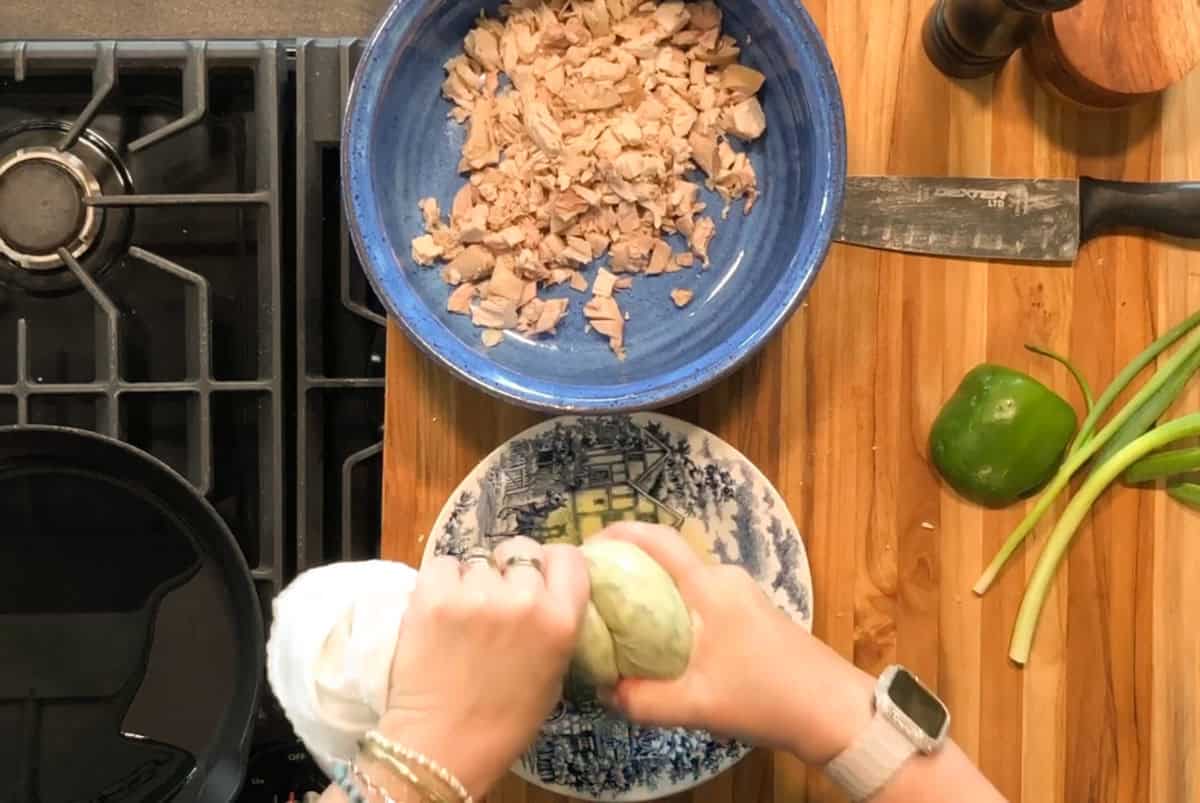 Overhead view of a person squeezing a cabbage over a patterned bowl. Next to them are a blue bowl with chopped tuna, a knife, a bell pepper, and green onions on a wooden counter beside a stovetop.