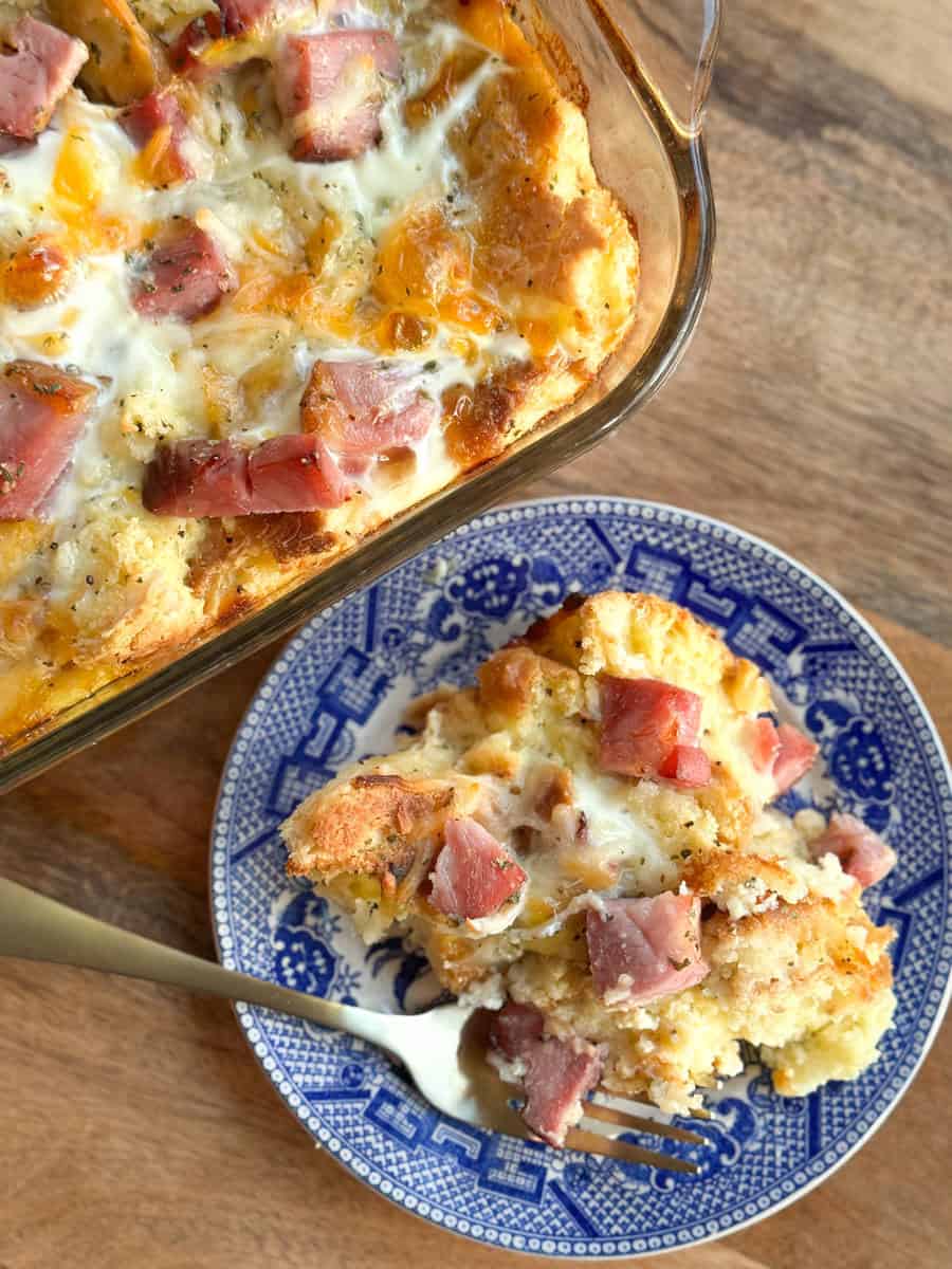 A slice of egg casserole with chunks of ham and melted cheese sits on a blue patterned plate next to a fork. The rest of the casserole is in a glass baking dish on a wooden surface.