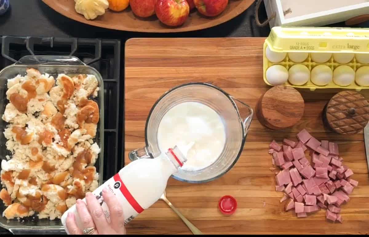 A hand pours milk into a glass bowl on a wooden counter with cubed ham, eggs in a carton, two round containers, a baking dish of torn bread, and a tray of apples and butter visible.