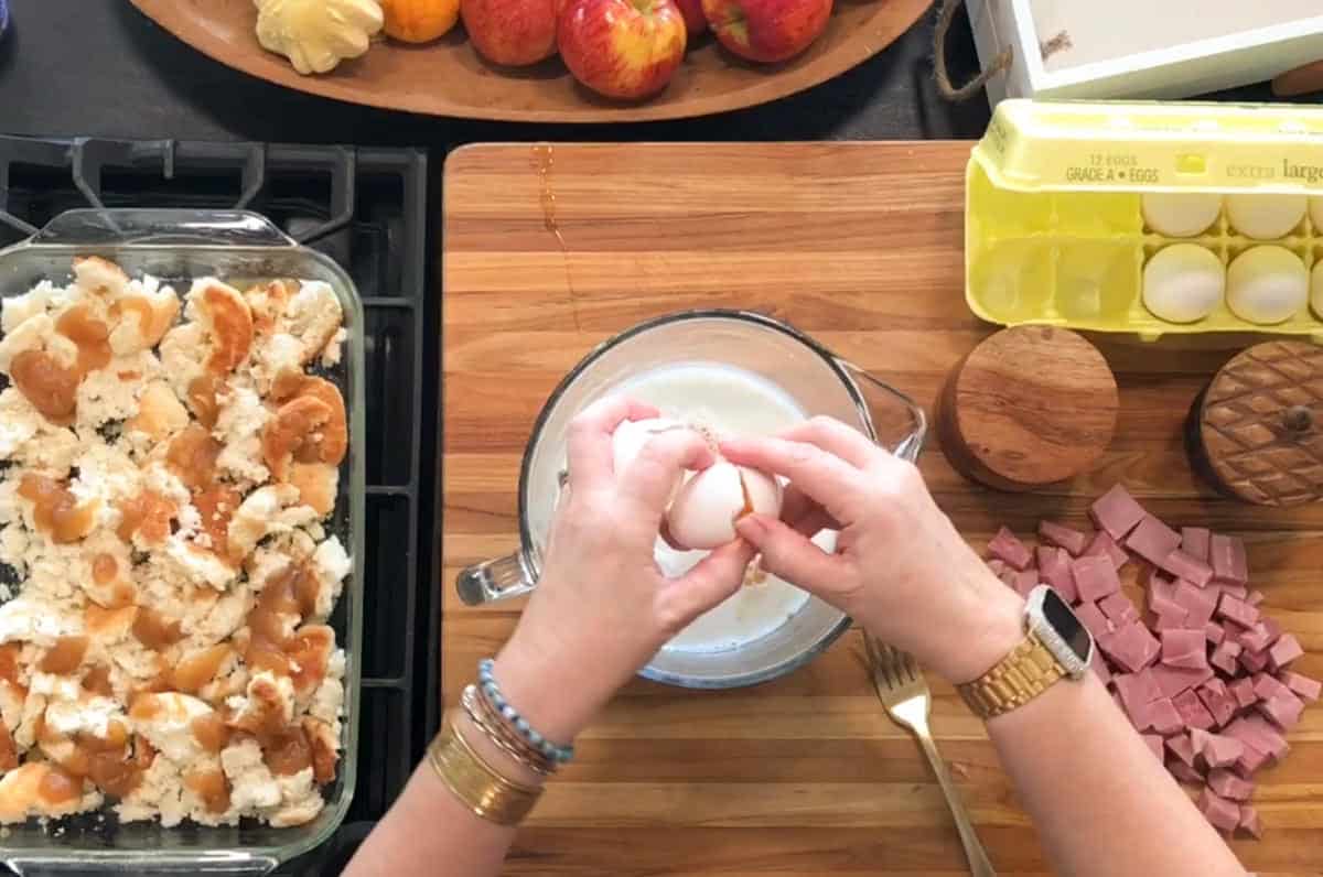 A person cracks an egg into a bowl of milk on a wooden countertop. Nearby are a dish of torn bread, a plate of apples, a carton of eggs, two wooden containers, chopped ham, and a fork.