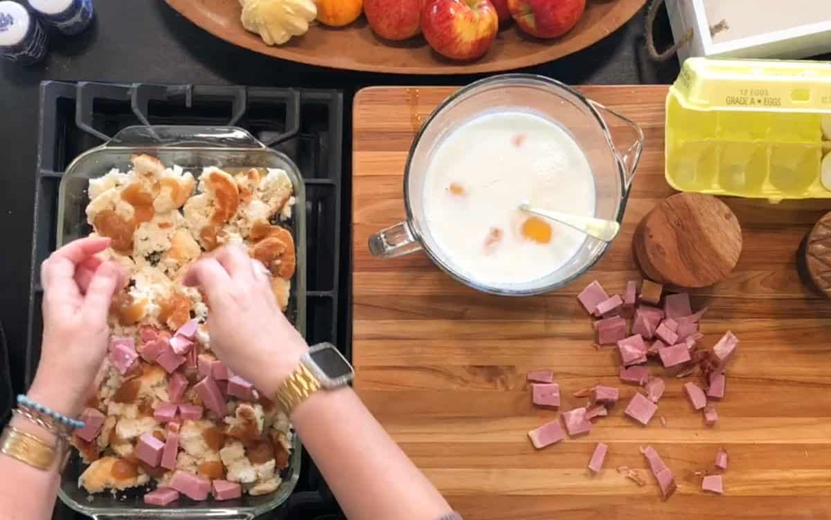 A person’s hands add cubed ham to a glass baking dish with torn bread pieces. Nearby are a bowl with milk and eggs, a cutting board, diced ham, egg carton, apples, and kitchen tools.