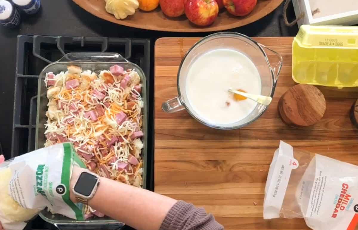 A person sprinkles shredded cheese over a casserole with bread and ham. Nearby are a bowl of eggs and milk being whisked, a wooden cutting board, eggs, apples, and kitchen utensils.