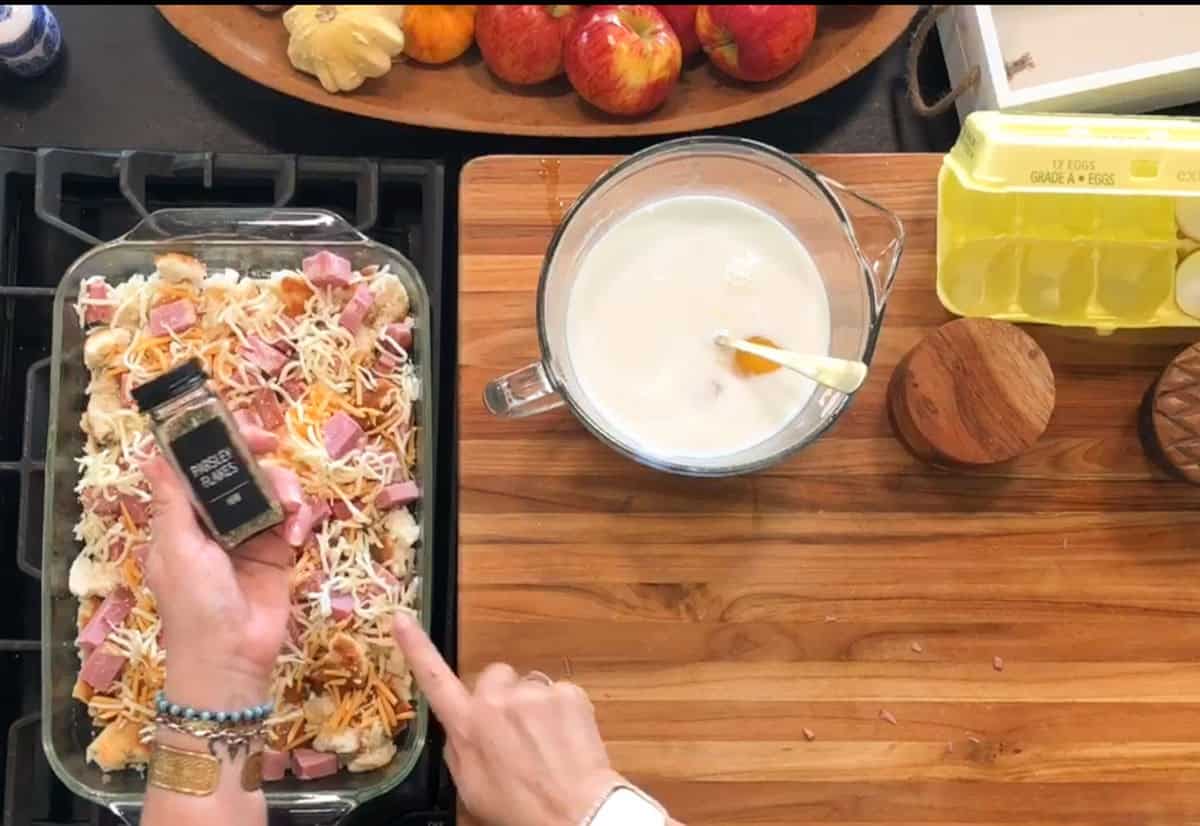 A person sprinkles black pepper onto a casserole dish filled with bread, ham, and shredded cheese. Next to it is a bowl of whisked eggs and milk, a carton of eggs, a wooden container, and a tray of apples.