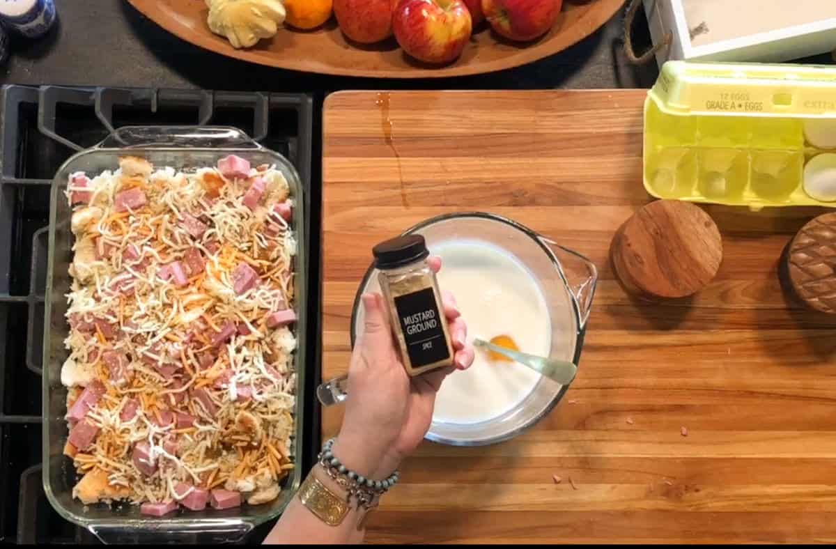 A hand holds a small container of ground mustard over a glass bowl of milk and eggs on a wooden counter. Next to it is a casserole dish with shredded cheese and ham, eggs in a carton, apples, and a wooden container.