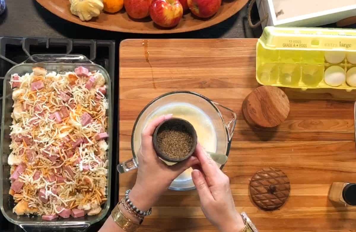 A person holds a small bowl of black pepper above a mixing bowl with a whisked mixture. Nearby are a dish of bread, ham, and cheese, a carton of eggs, wooden lids, and apples on a wooden cutting board.
