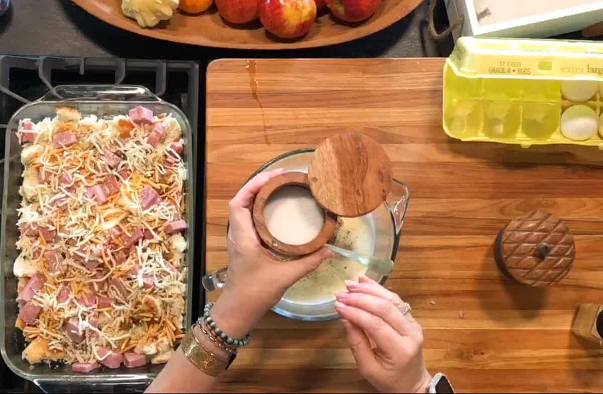 A person adds salt to a measuring cup with eggs and milk on a wooden cutting board; a baking dish with a layered casserole and a carton of eggs are nearby.