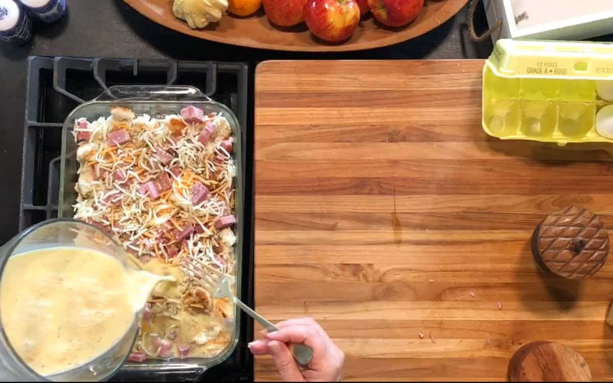 A person pours beaten eggs from a glass pitcher onto a casserole with bread, ham, and shredded cheese in a glass baking dish on a stovetop. An empty egg carton, a wooden bowl, and apples are on a wooden counter nearby.
