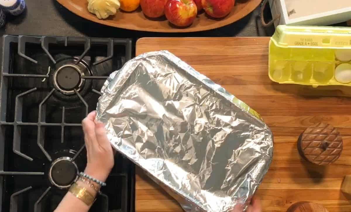 Two hands are placing a foil-covered baking dish on a wooden countertop next to a carton of eggs, a wooden container, and a tray of apples. A stovetop with burners is visible on the left side of the image.