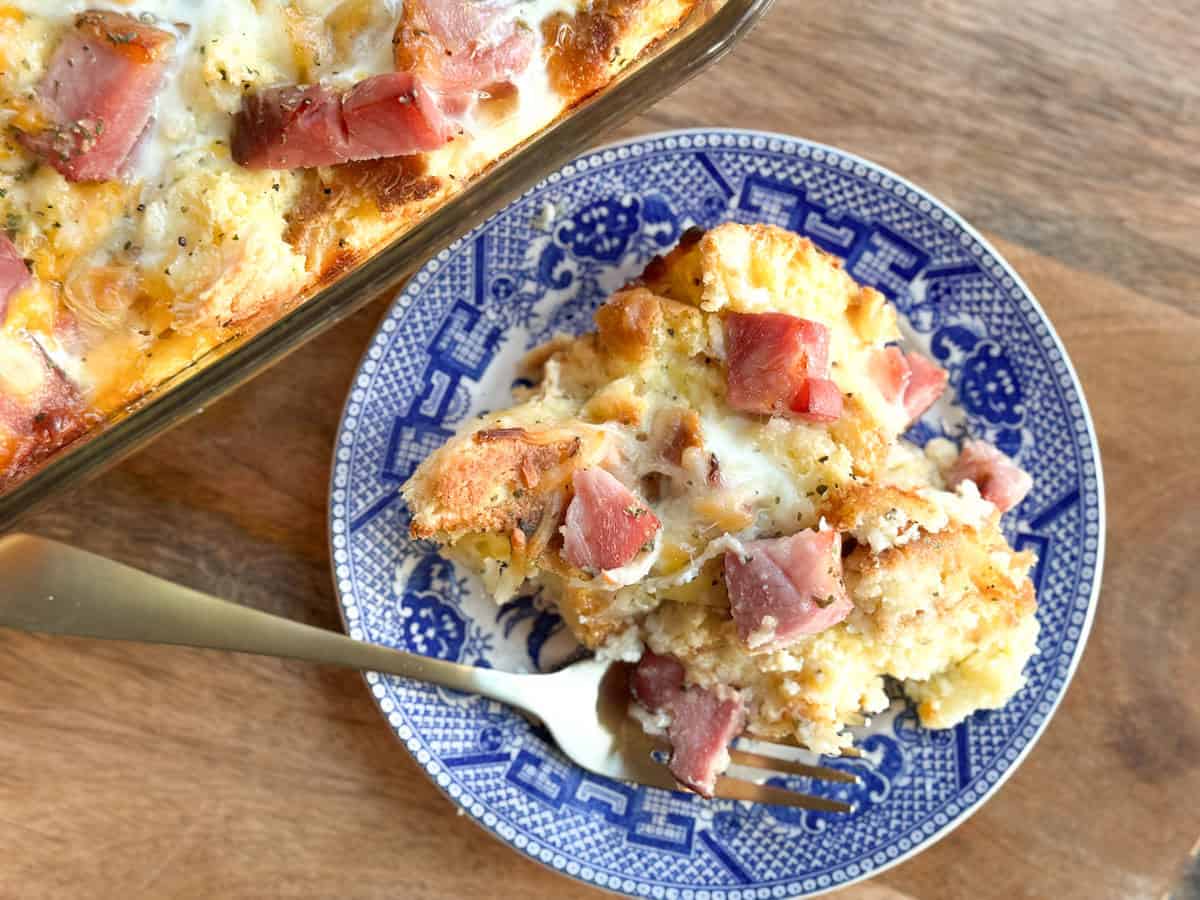 A blue and white plate holds a serving of egg casserole with chunks of bread, cheese, and ham. A fork rests on the plate. A larger casserole dish with the same food is partially visible on a wooden surface.