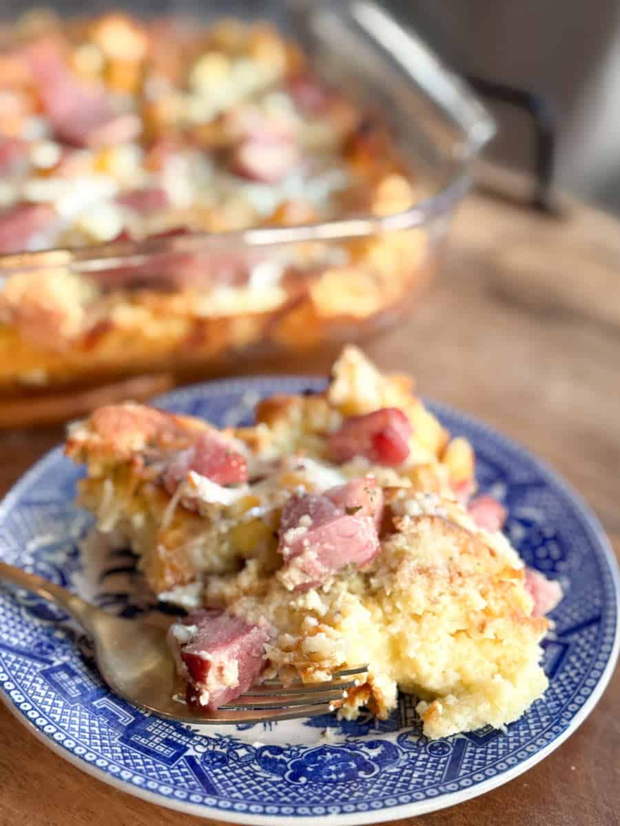 A close-up of a plate with a serving of ham and cheese breakfast casserole, with a fork beside it. In the background, a glass baking dish with more casserole is slightly out of focus.