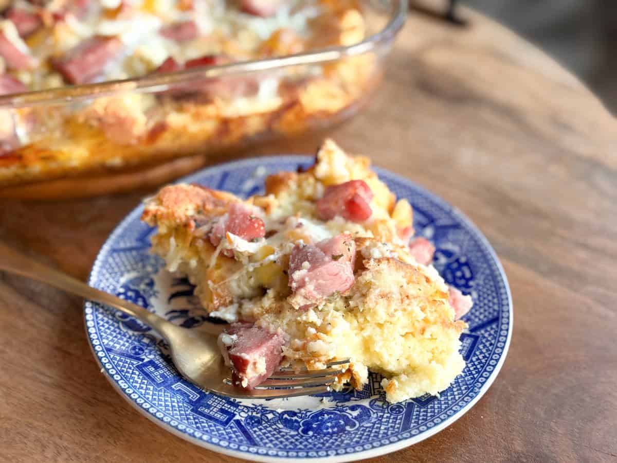 A slice of casserole with visible chunks of ham and melted cheese is served on a blue and white plate with a fork. The rest of the casserole is in a glass baking dish in the background.
