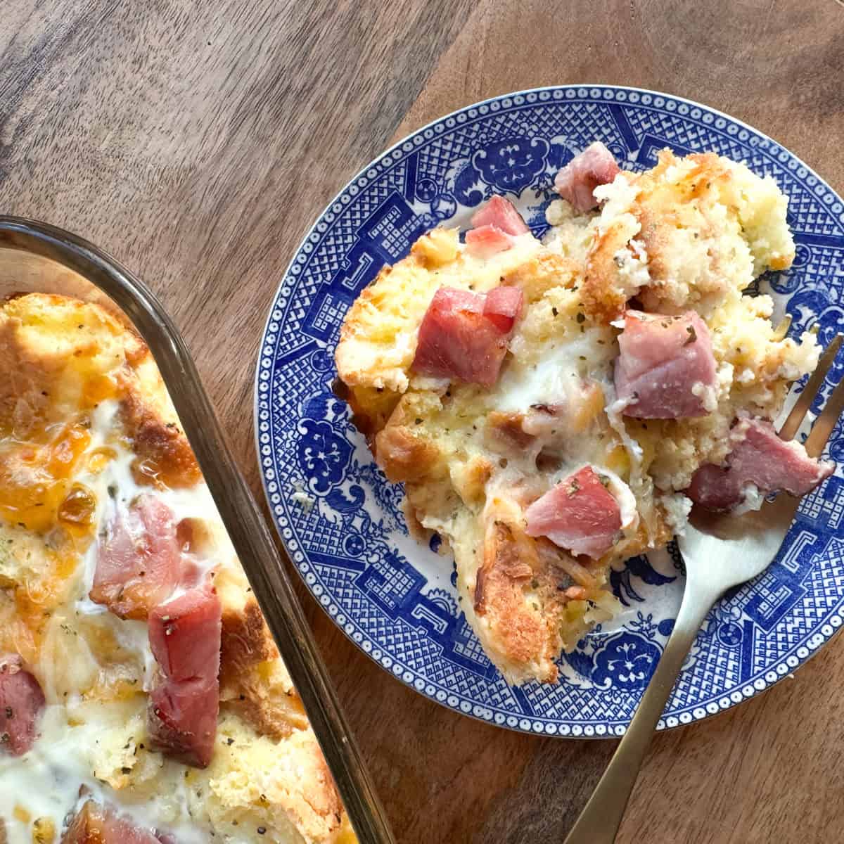 A serving of egg, bread, and ham casserole is plated on a blue and white patterned dish with a fork. The remaining casserole is visible in a glass baking dish beside the plate on a wooden surface.