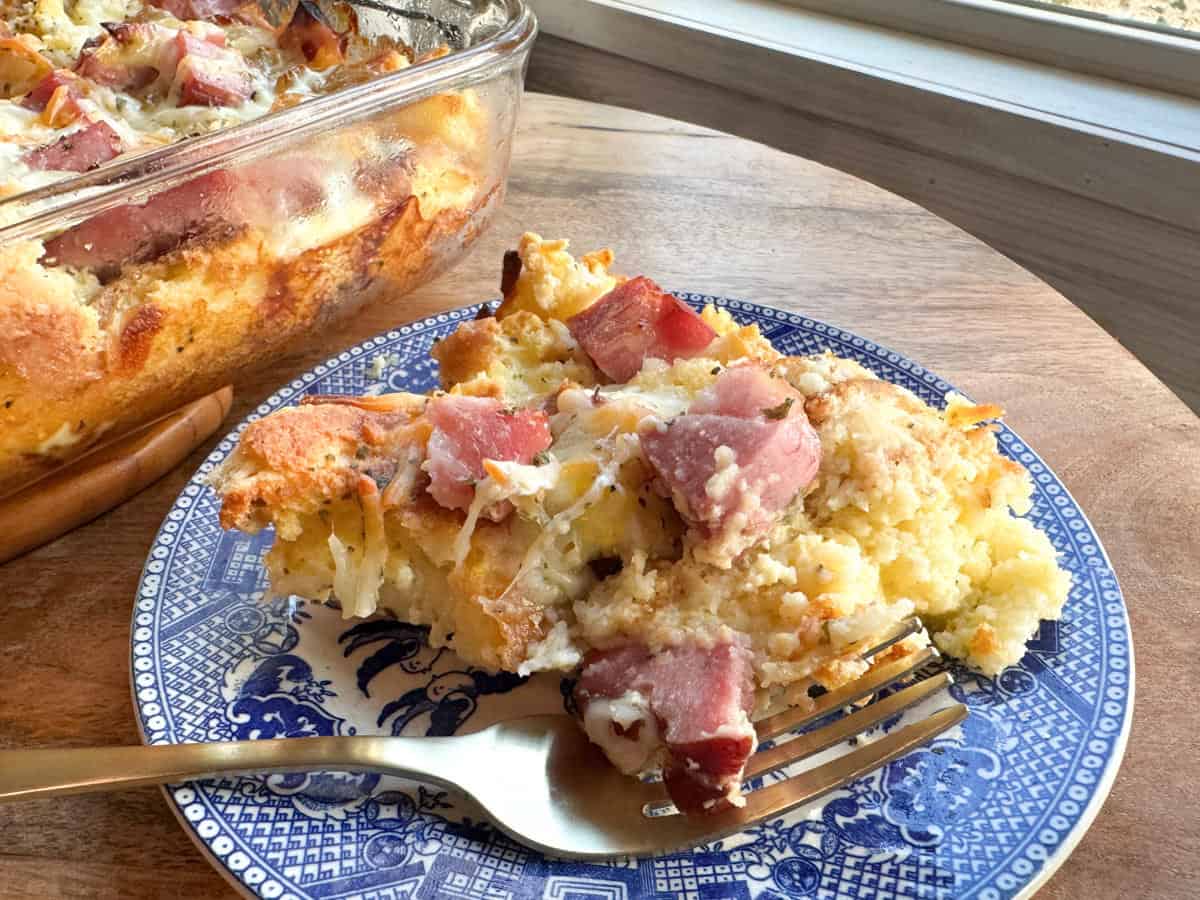 A slice of casserole with visible chunks of ham and melted cheese sits on a blue and white plate next to a fork. The casserole dish with more servings is in the background on a wooden surface by a window.