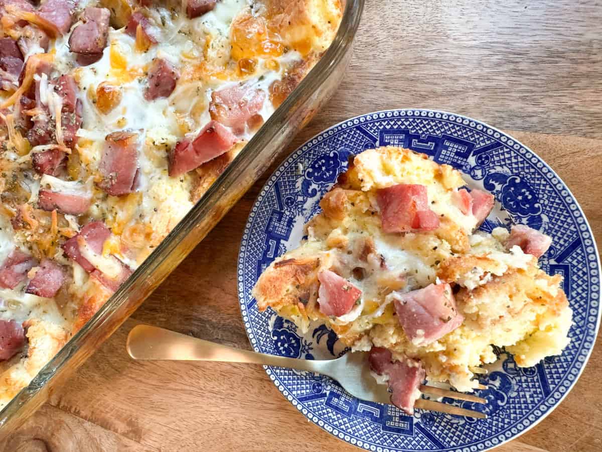 A serving of ham and cheese breakfast casserole on a blue and white plate sits next to a glass baking dish with the remaining casserole. A fork rests on the plate atop a wooden surface.
