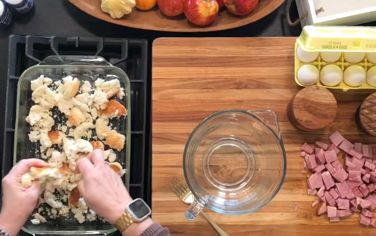 A person tears bread into pieces in a glass baking dish on a wooden countertop next to a bowl, a fork, chopped ham, eggs in a carton, a wooden container, and a tray of apples.