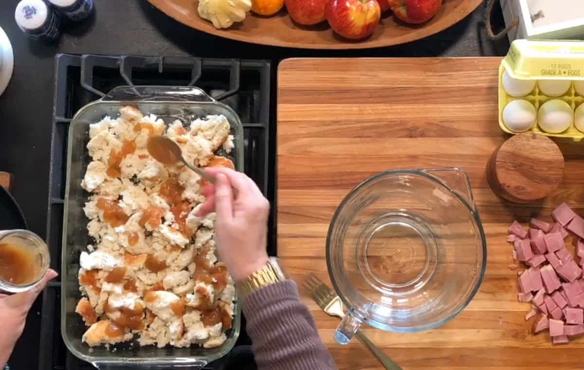 A person spoons liquid over torn pieces of bread in a glass baking dish on a stove. Nearby are a mixing bowl, eggs, cubed ham, apples, and kitchen utensils on a wooden cutting board.