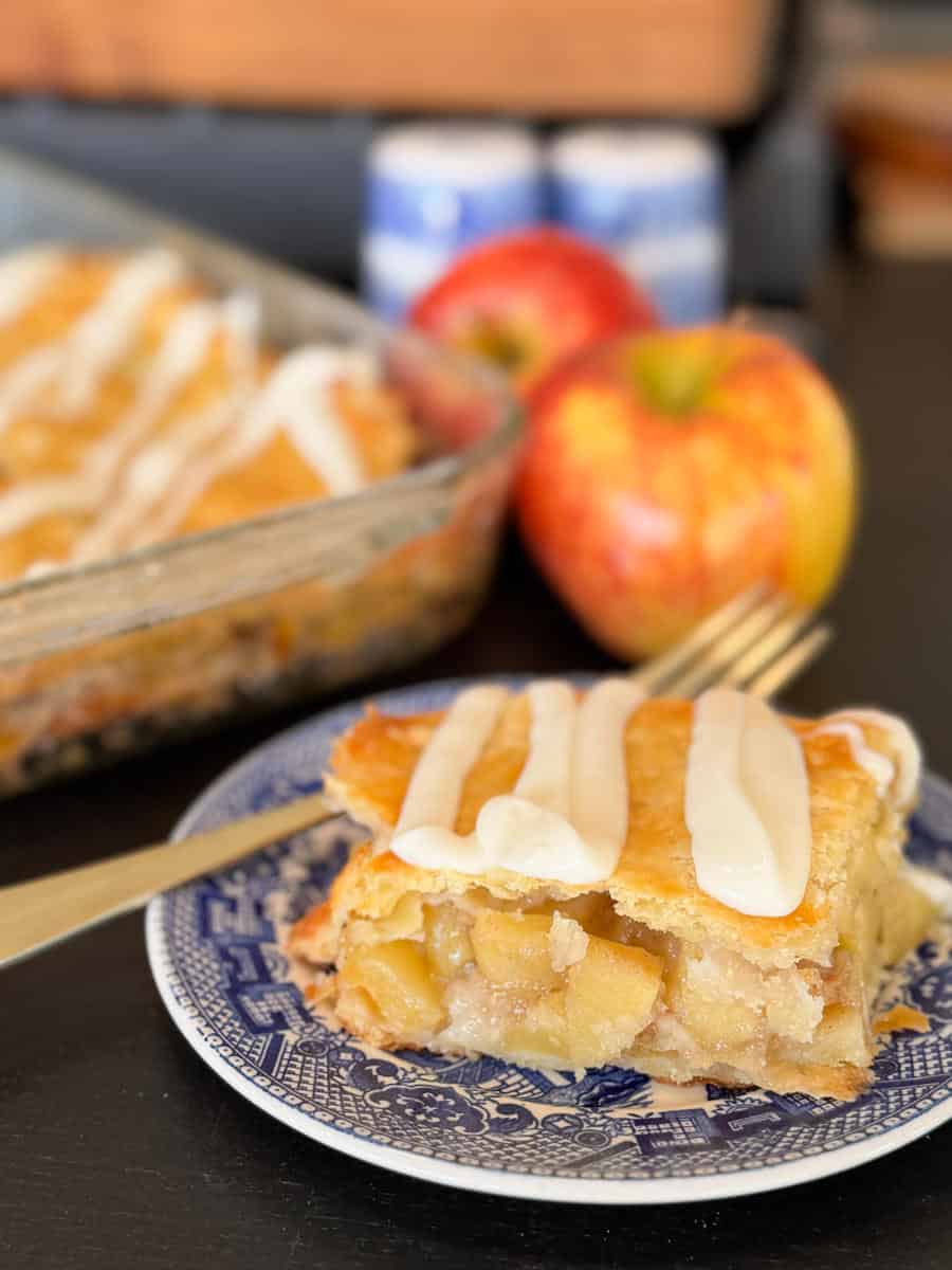 A slice of apple pie with icing on top sits on a blue and white plate with a fork. In the background, there is a baking dish of pie, two red apples, and two small blue and white jars.