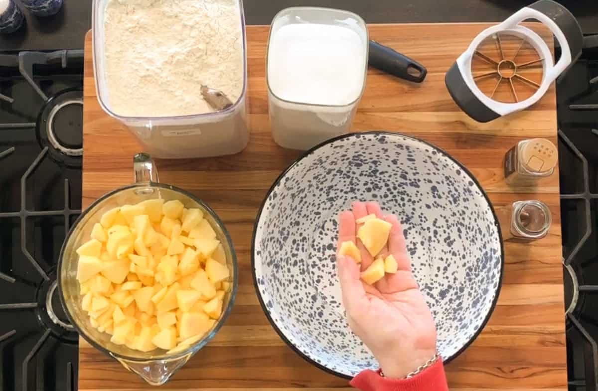 A hand holds pieces of chopped apples over an empty speckled mixing bowl on a wooden surface, surrounded by containers of flour, sugar, a bowl of more chopped apples, an apple slicer, and a spice jar.
