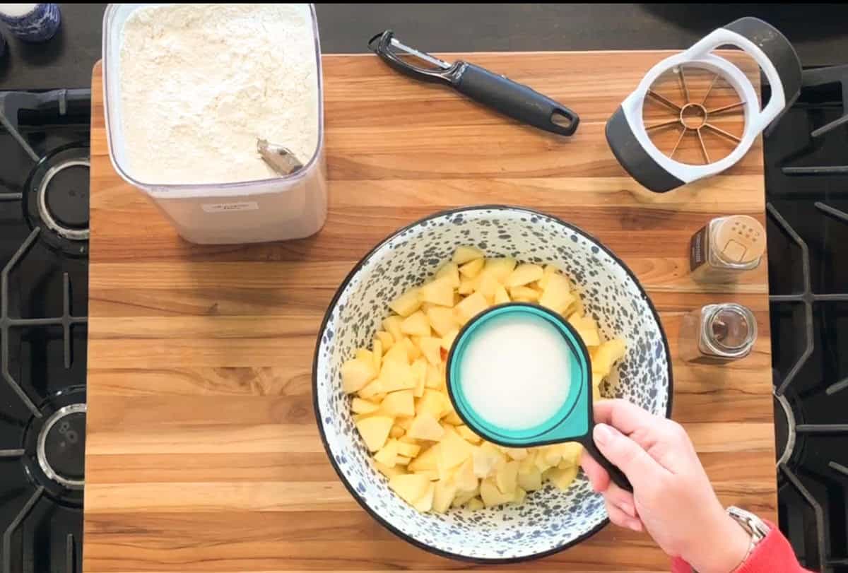 A hand pours sugar from a green measuring cup into a bowl of chopped apples. Nearby are a flour container with a scoop, a peeler, an apple slicer, and jars of cinnamon and nutmeg on a wooden countertop.