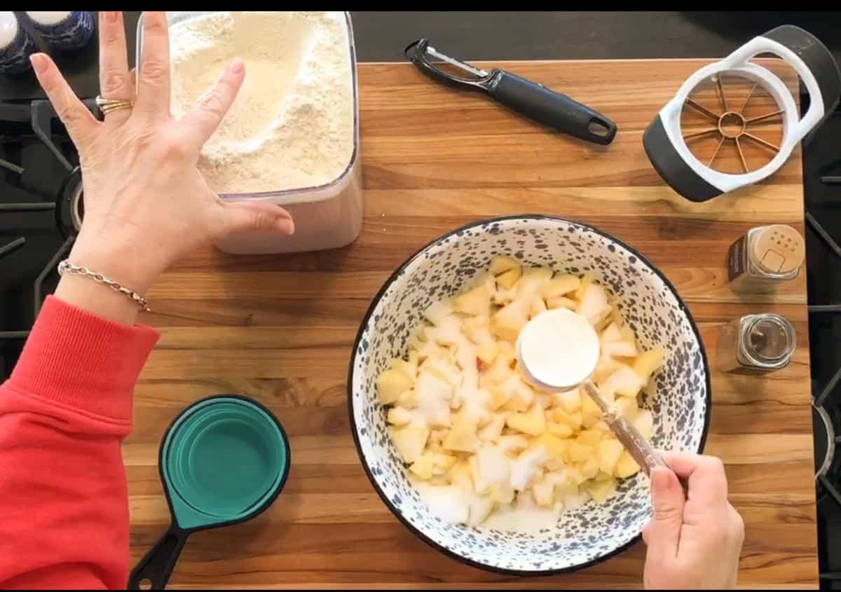 A person prepares an apple mixture in a bowl with chopped apples and granulated sugar, while holding a container of flour. Measuring cups, a peeler, an apple slicer, and spices are on a wooden countertop.